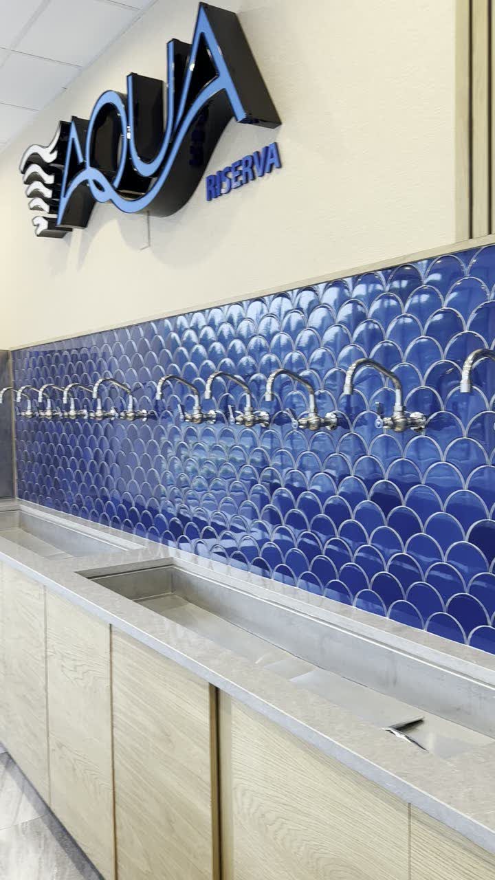 The Aqua Riserva tap wall — ten dispensing taps over blue fish-scale tile above a stainless drain trough